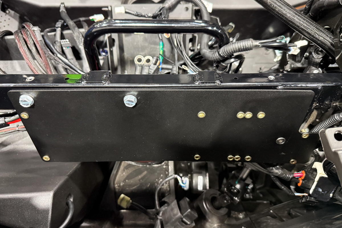 Black metal mounting plate installed on vehicle chassis with visible bolts and wiring harness