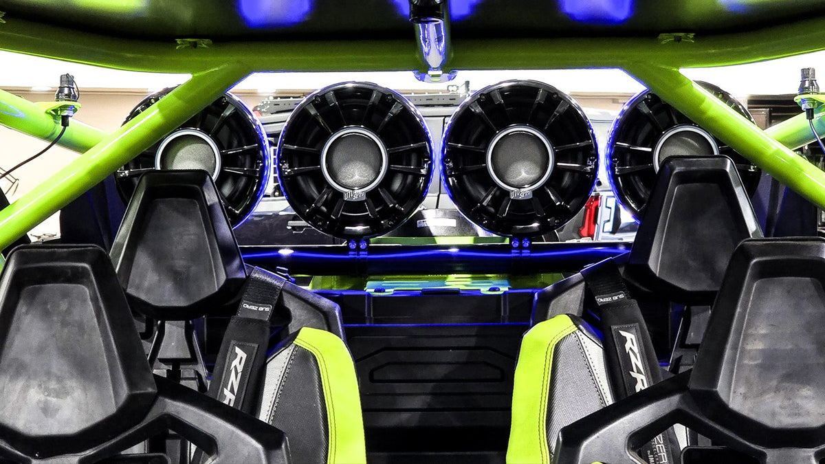 Four circular UTV Stereo speakers mounted on a green roll cage behind black bucket seats